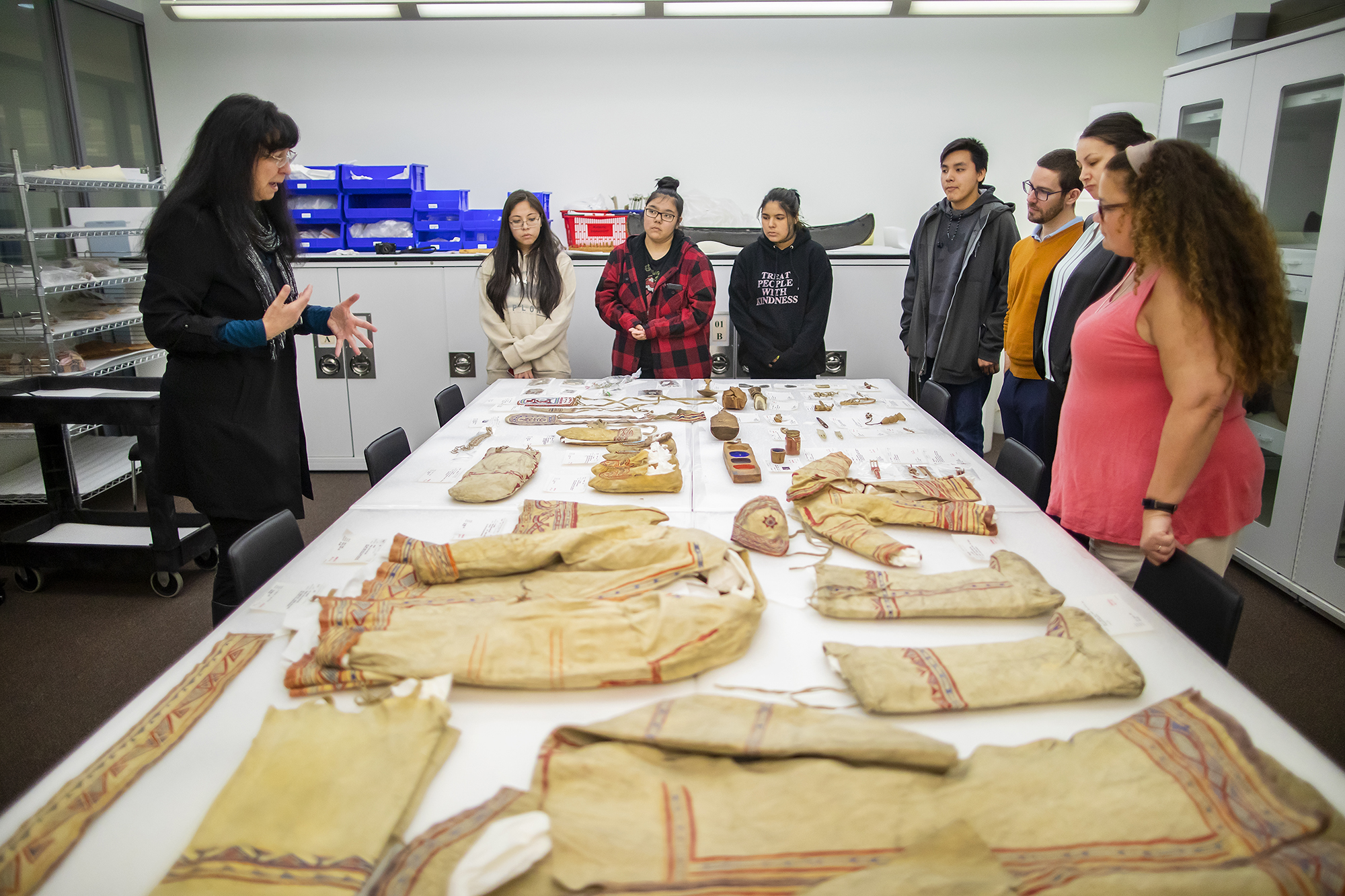 Professor stands on one side of a table filled with Native American objects while talking to seven other people across from and beside her i