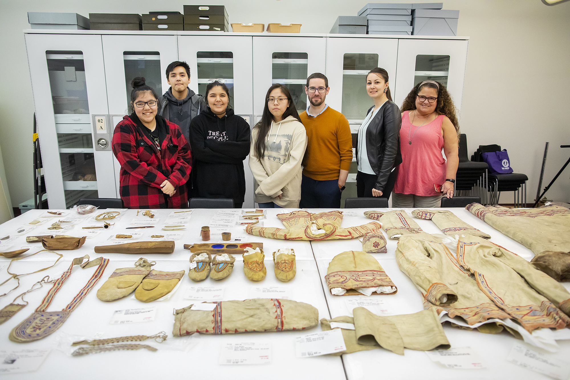 The seven visitors from the Naskapi Nation group standing in front of table filled with Native American objects. 