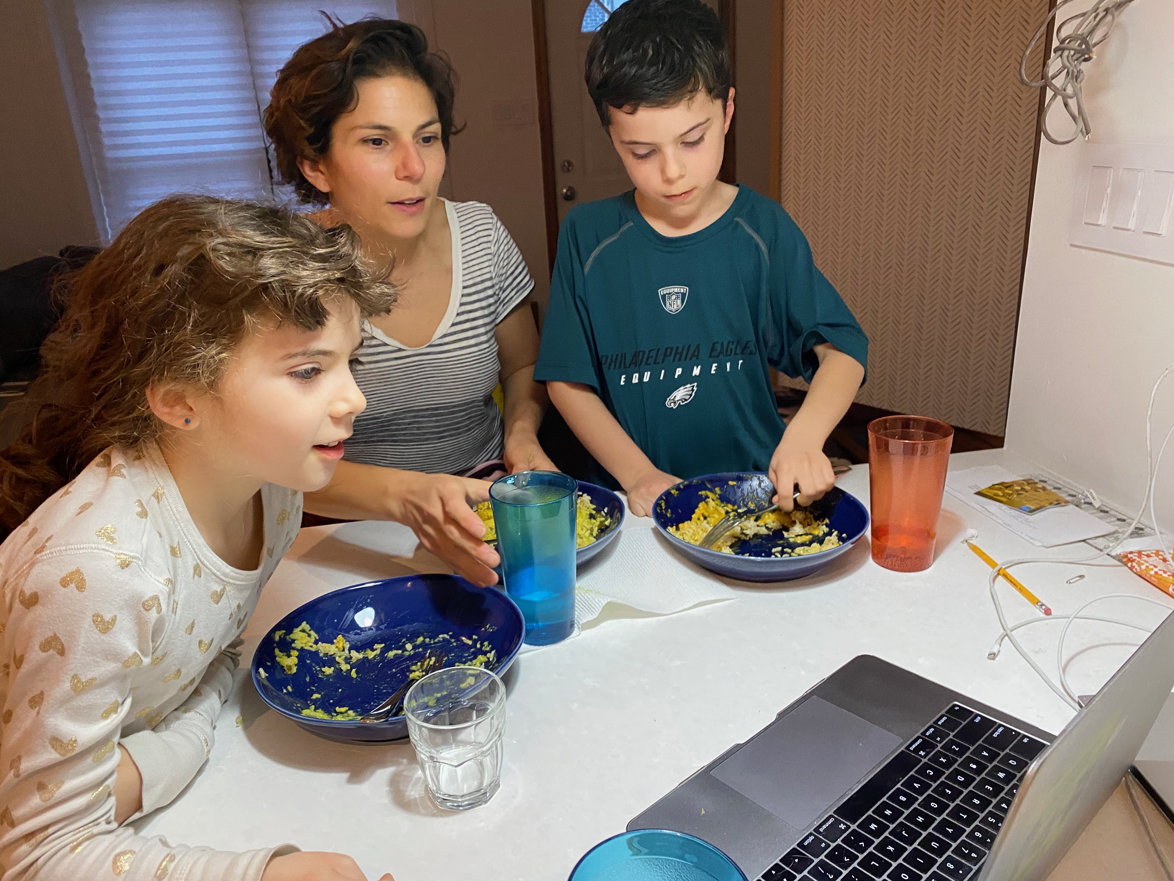 A professor and two children eating from bowls at a kitchen counter with a laptop computer open in front of them. 