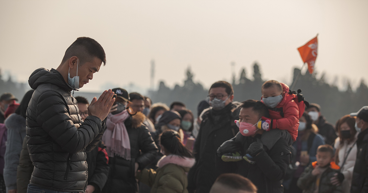 Person praying in front of a crowd in China with mask down around their chin, people wearing masks walk around them, outside a temple.