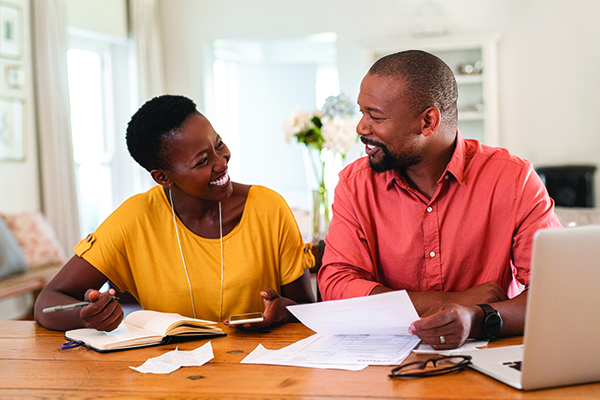 A couple sits at a desk in their living room smiling at each other while holding papers and discussing health care coverage