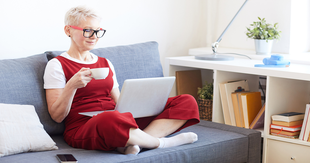 Person sitting cross-legged on a couch holding a tea cup looking at a laptop