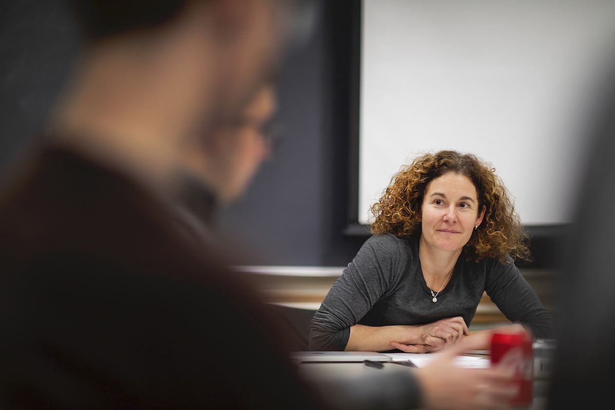 Person sits at desk in front of chalkboard with hands clasped, looking at another person speaking.