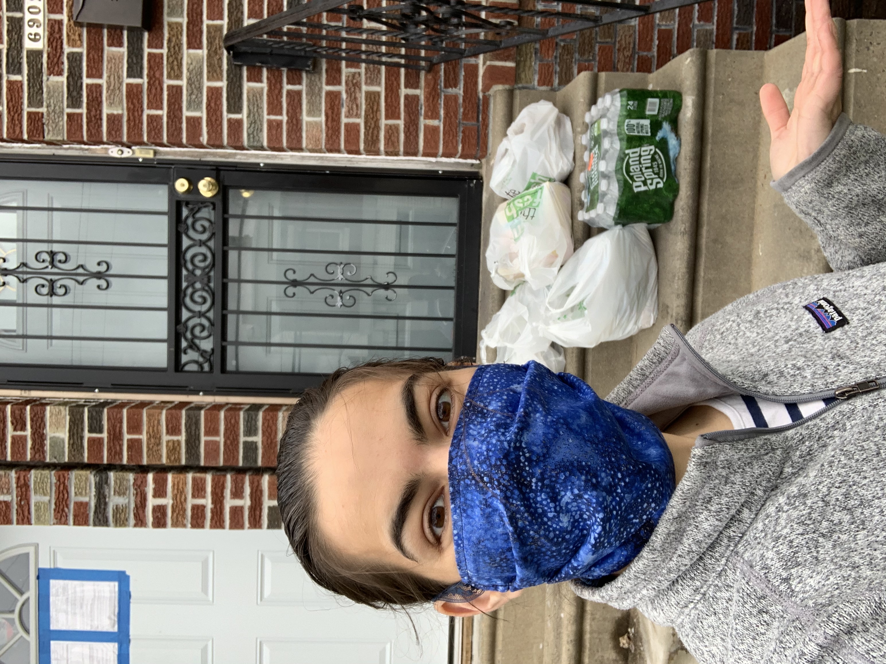 A person in a fleece and face mask in front of a stoop that has a 24-pack of bottled water and four plastic bags full of groceries.