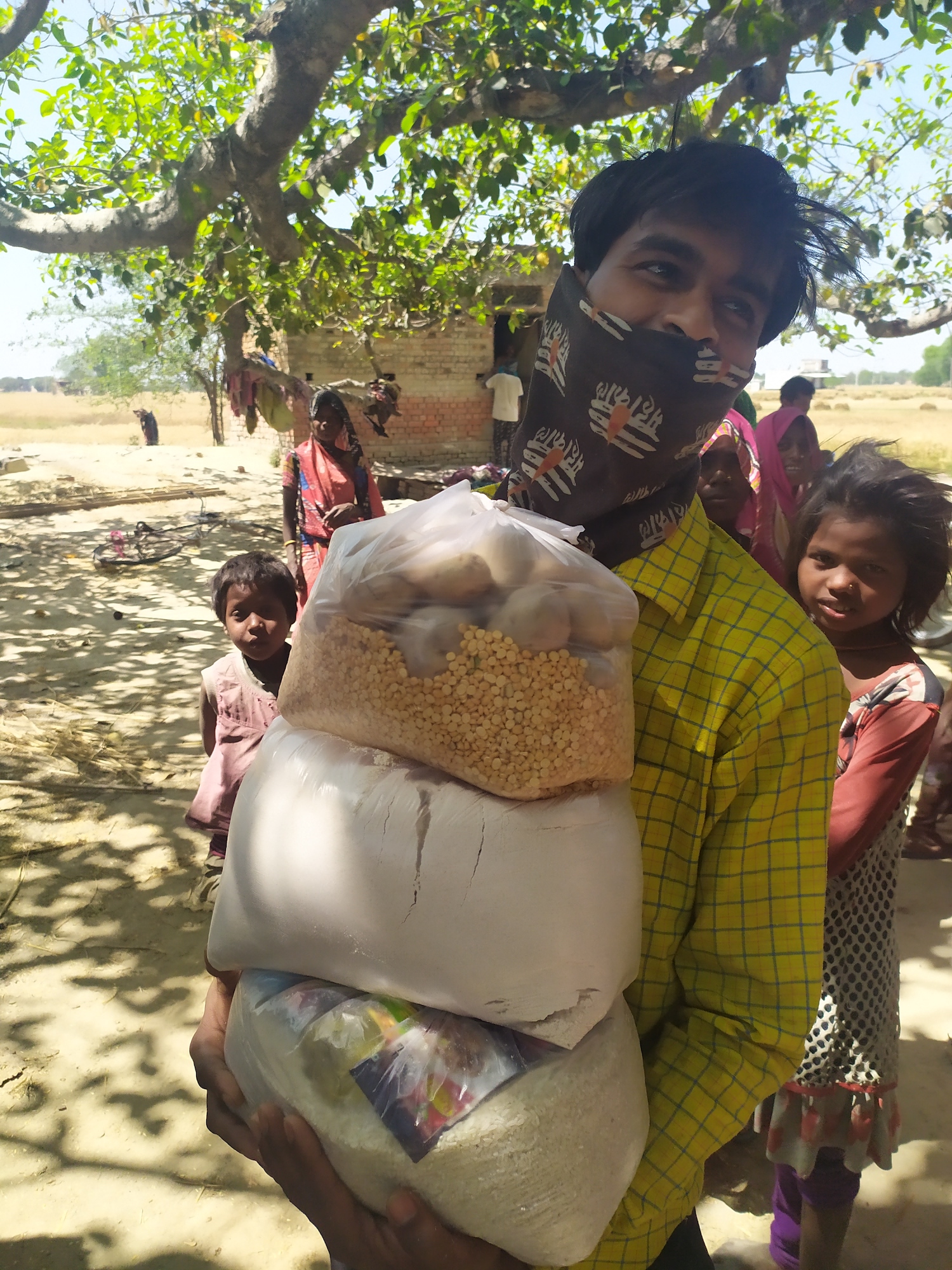 Person with bandana over face holds a bag of food and smiles with children in backgroud