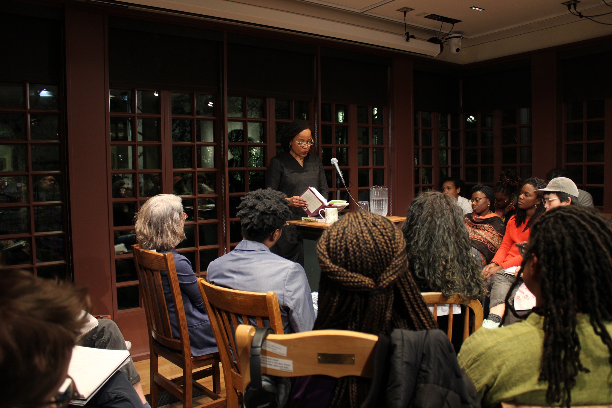 Writer reading from a book with 10 people seated in front of her in a windowed room. 