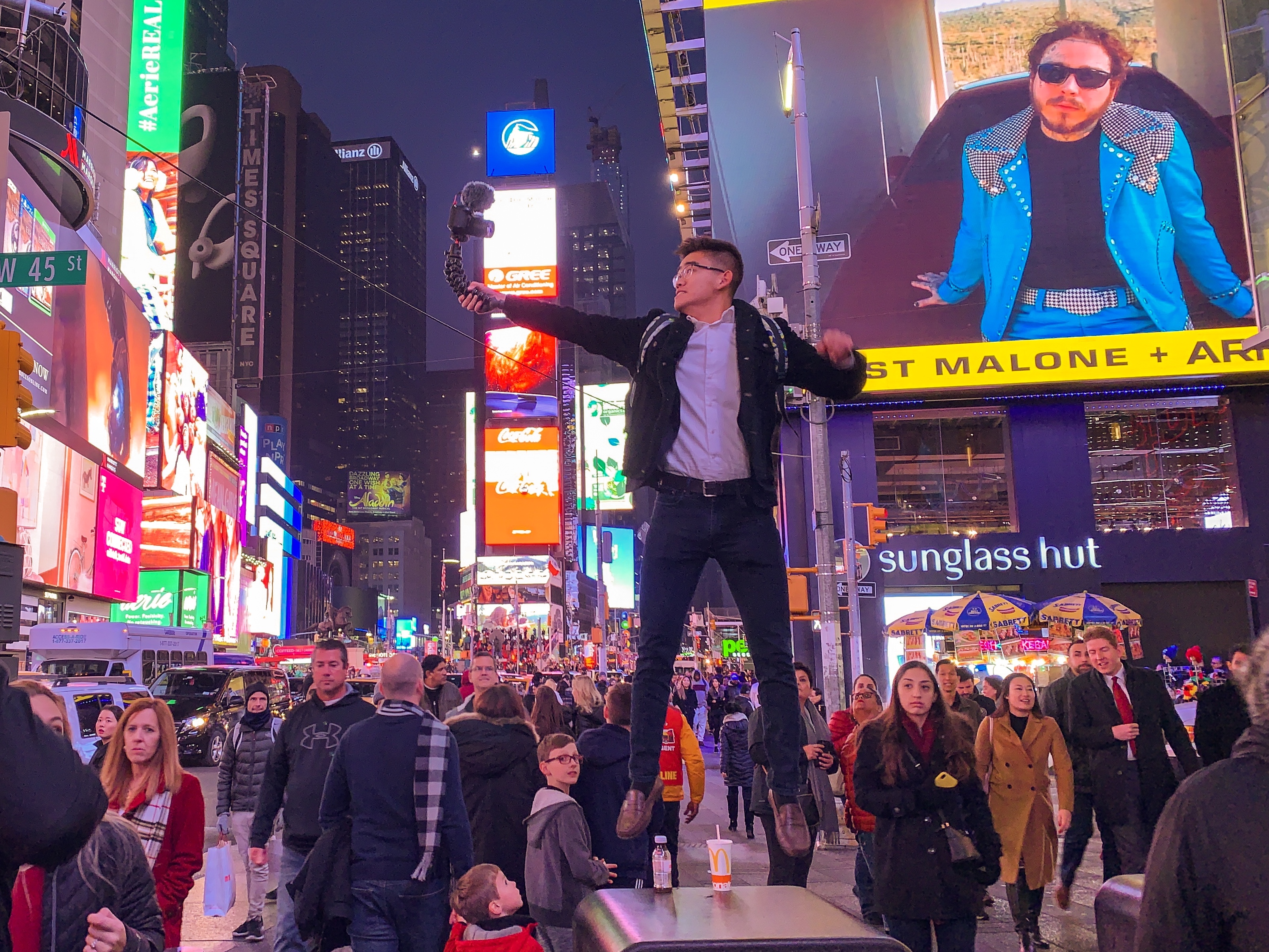 Young man holding a camera jumps above a trash can in the middle of busy Times Square