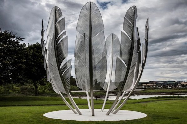 A large stainless steel sculpture depicting bird feathers standing upright in a circle sits in a green grass field.