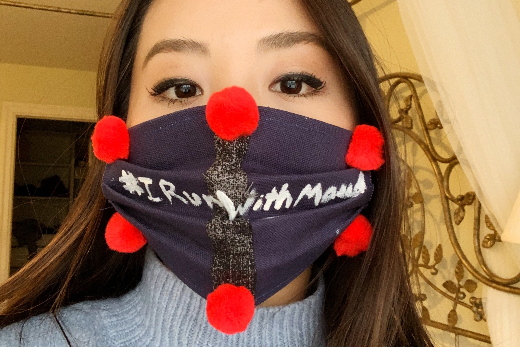 student wearing a dark mask with pom-poms with words #IRunWithMaud