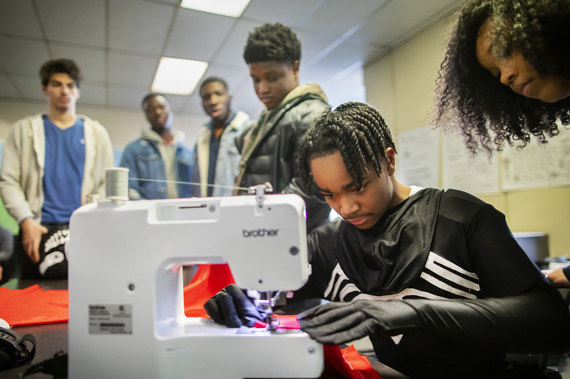 Costumed student feeds cloth through a sewing machine as five others look on