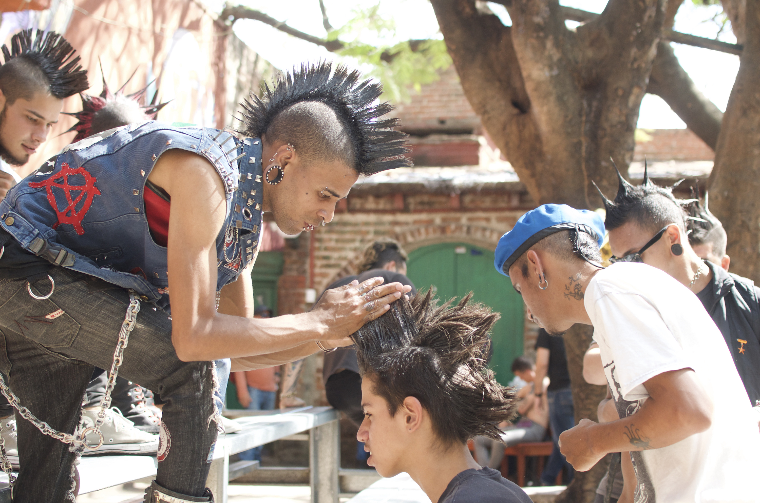 A young mohawked man with a leather vest featuring a red anarchy symbol styles another young man's hair into a mohawk