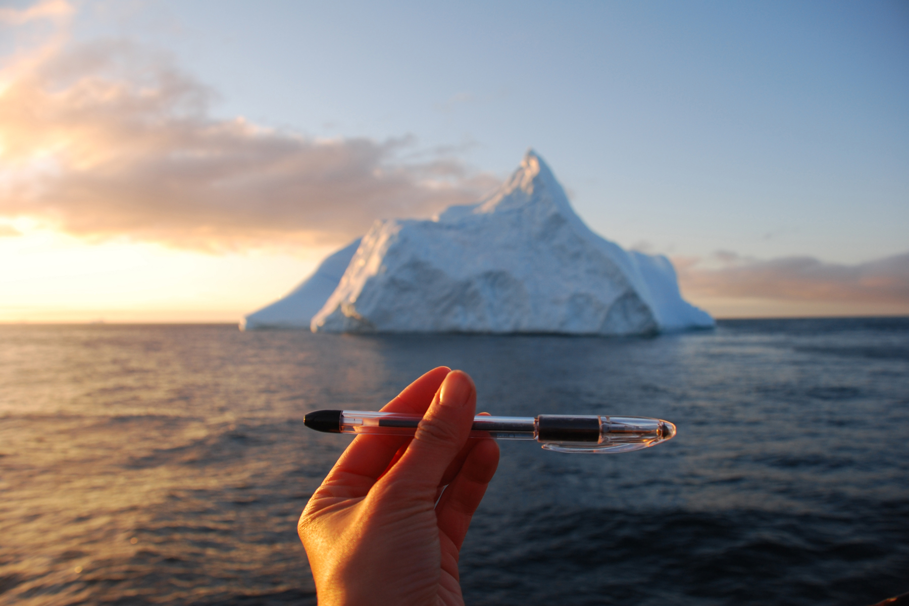 A hand holds a pen in front of an iceberg in the ocean