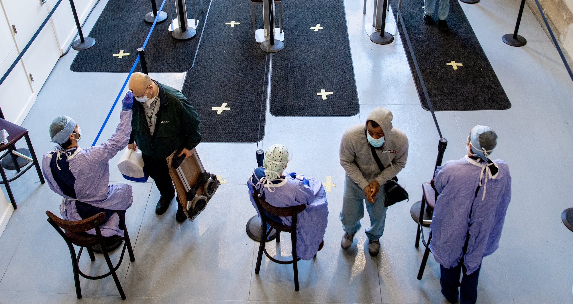 The entrance to a hospital. People in personal protective equipment swab others as they enter the building.