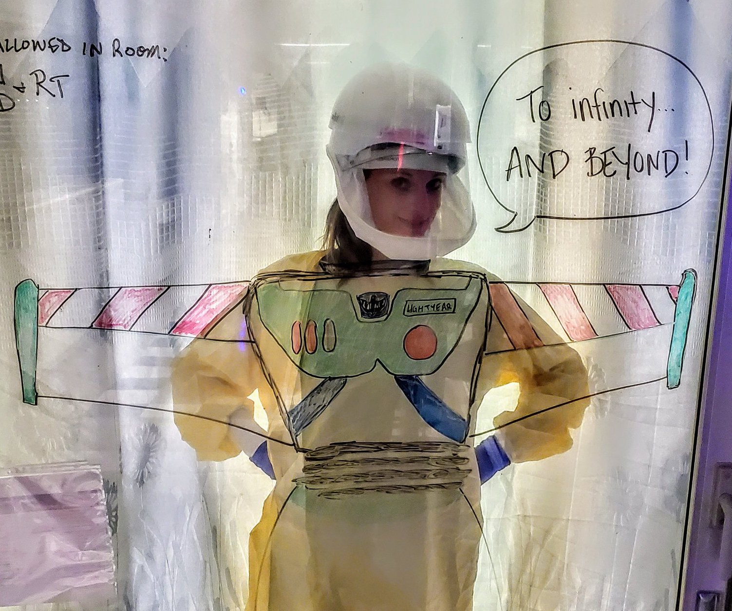 A person standing behind a glass wall wearing personal protective equipment. On the glass is a drawing of the cartoon character, Buzz Lightyear, with the words, "To infinity and beyond..."