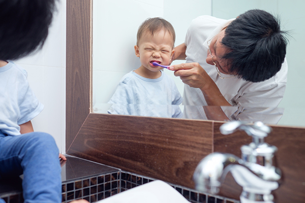 Parent brushes their toddler’s teeth seen in a bathroom mirror