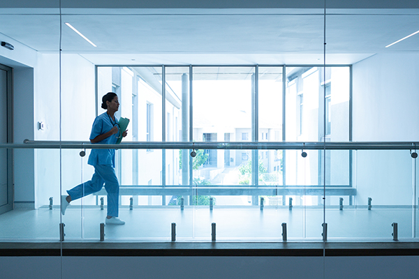 A nurse holding paperwork and wearing a stethoscope runs through a hallway past glass windows
