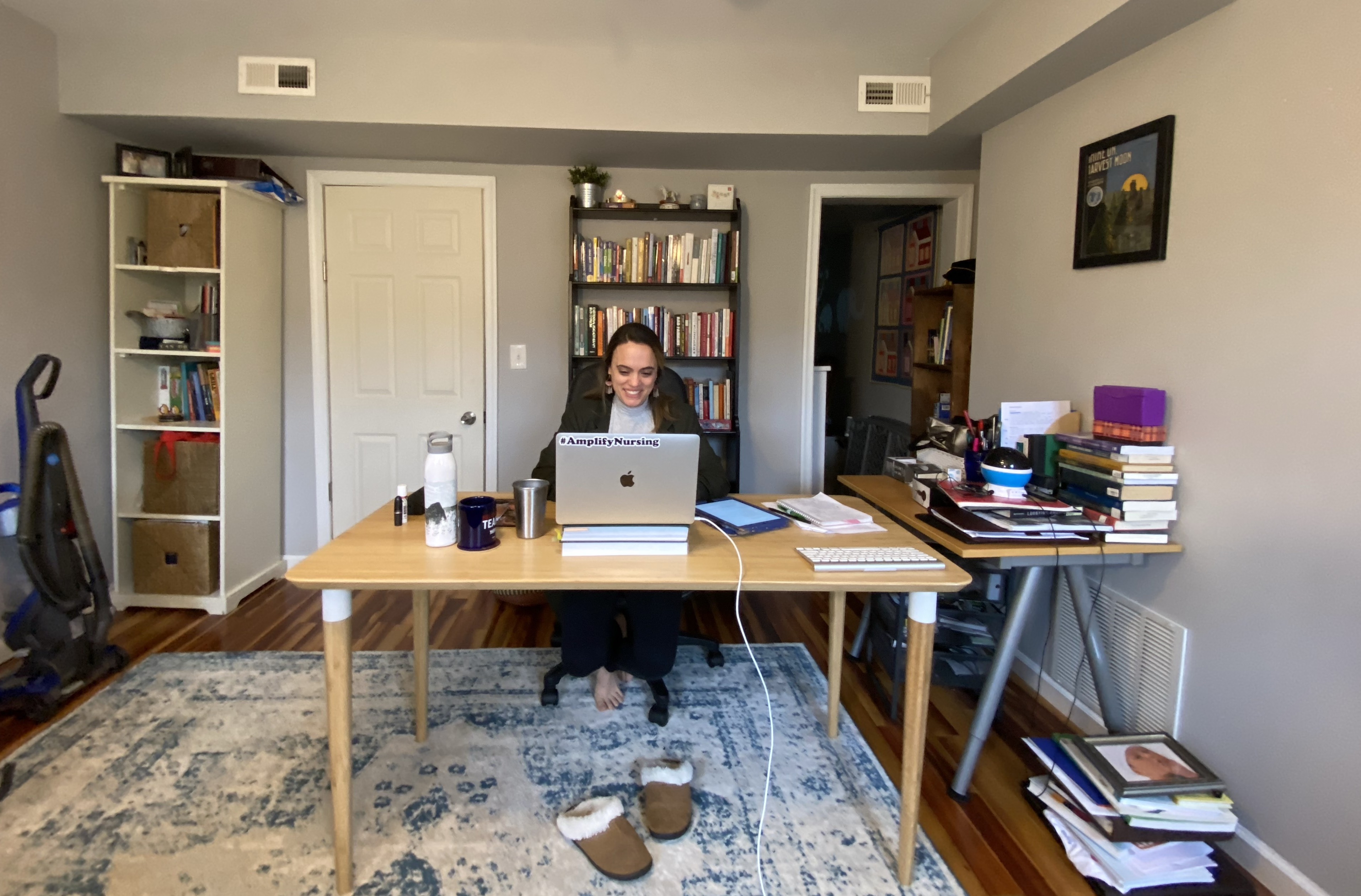 A person sitting at a desk behind a computer. The room has books piled up on the desk, and two bookshelves in the background, along with a vacuum to the side and slippers on the floor in front.