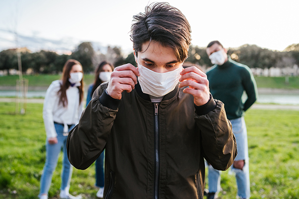 A teenager stands outside and puts a mask on their face while a group of friends wearing masks stands behind them on the grass.