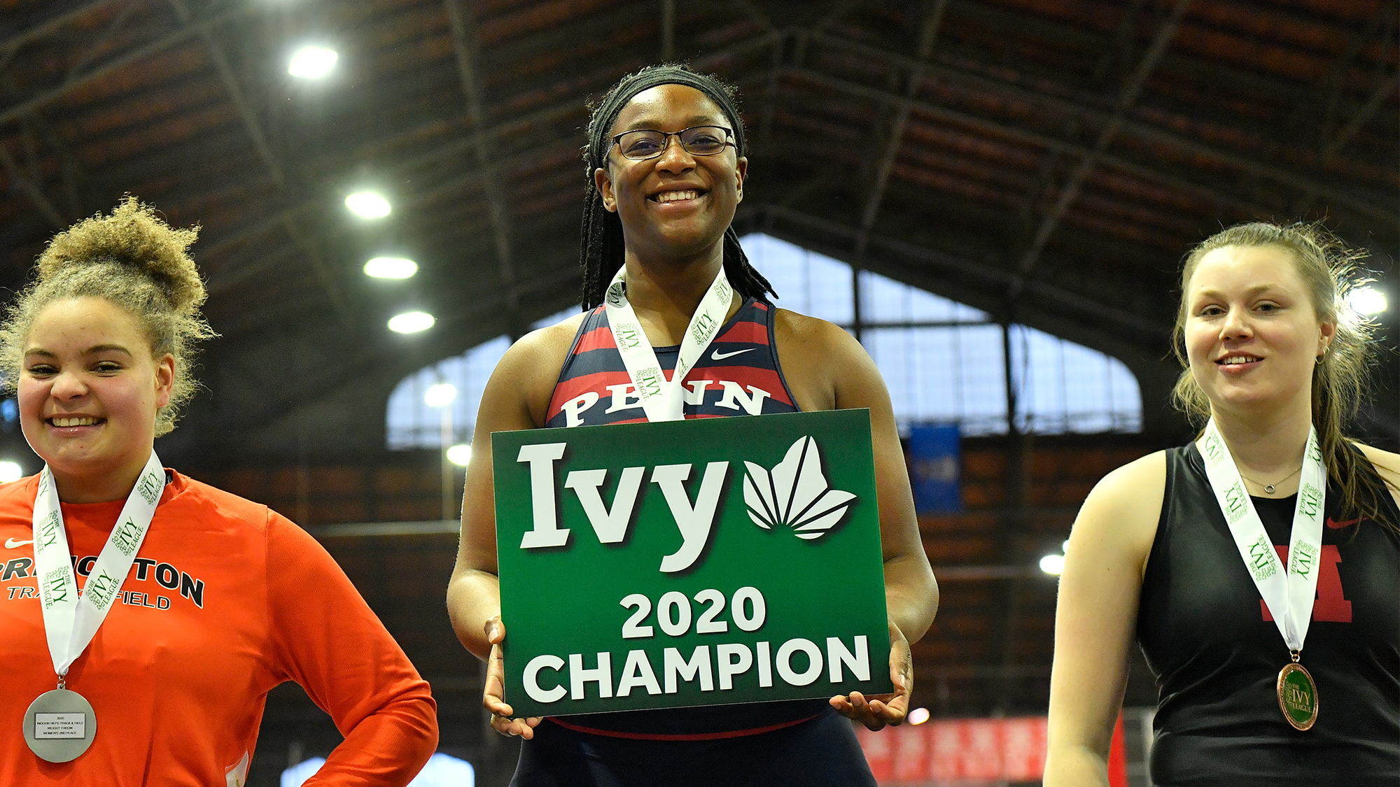 Mayyi Mahama stands on the first place podium at the 2020 Indoor Ivy Heps, holding a first place board.