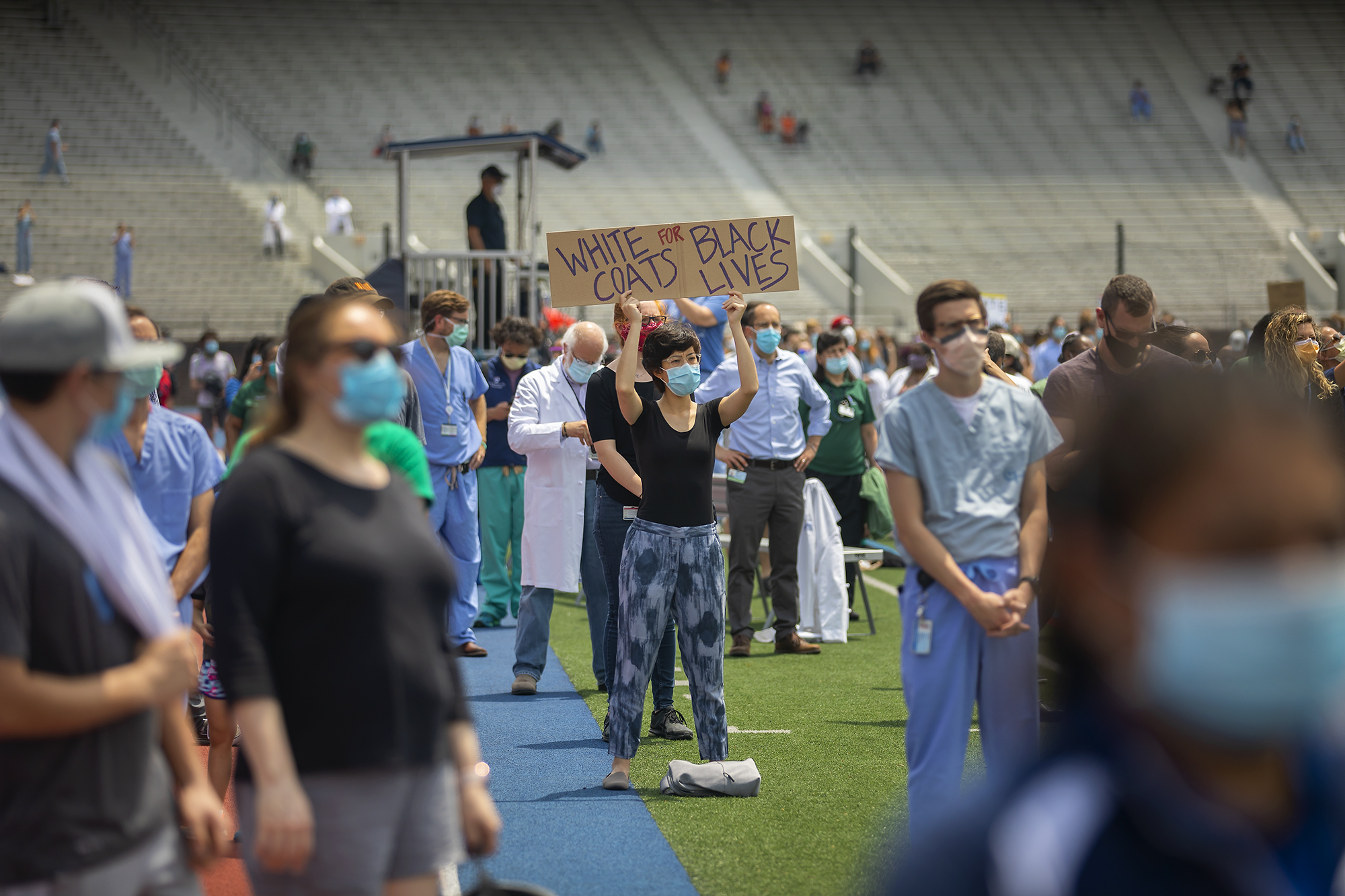 A crowd of health care personnel wearing protective face masks stand on Franklin Field in support of Black Lives Matter, one person holds a sign that reads White Coats for Black Lives.