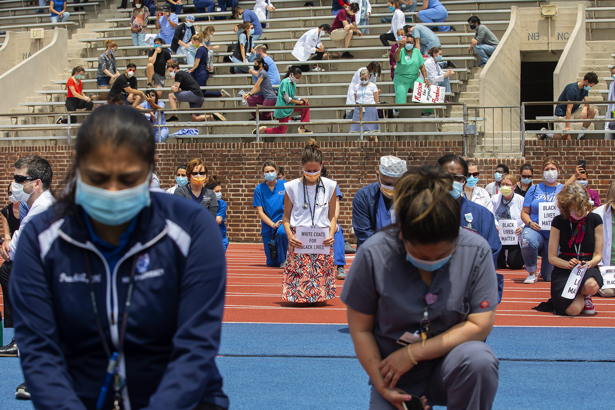 Penn Medicine workers in scrubs and protective face masks kneel on Franklin Field, one holds a sign that reads White Coats for Black Lives