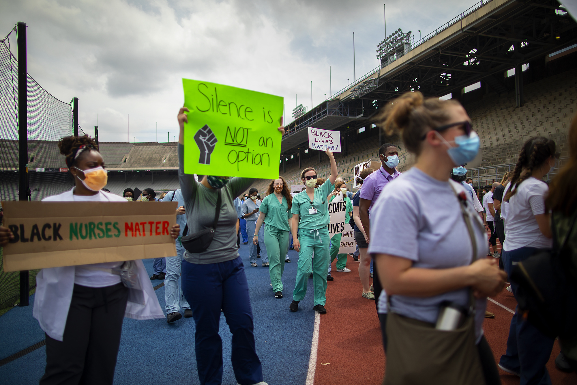 Medical personnel in scrubs, white coats and face masks on the track at Franklin Field, one holds a sign reading Black Nurses Matter, another holds a sign that reads Silence is NOT an option with a raised fist image next to the words.