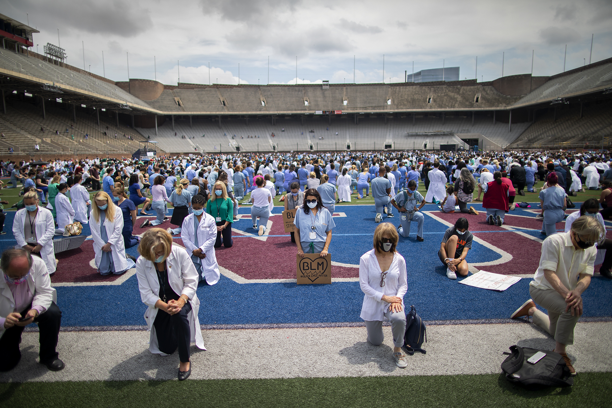 Medical personnel in scrubs, white coats and face masks kneel on the field at Franklin Field, one holds a sign with the letters BLM surrounded by a heart.