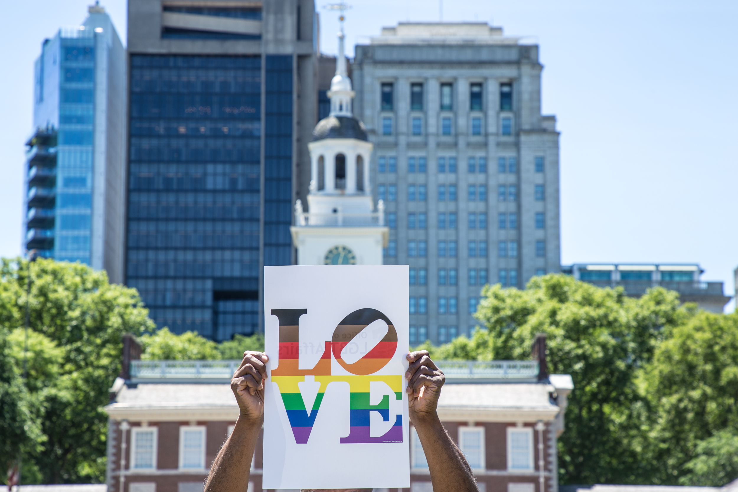 A sign with LOVE written the colors of the black and brown rainbow flag is held aloft before City Hall