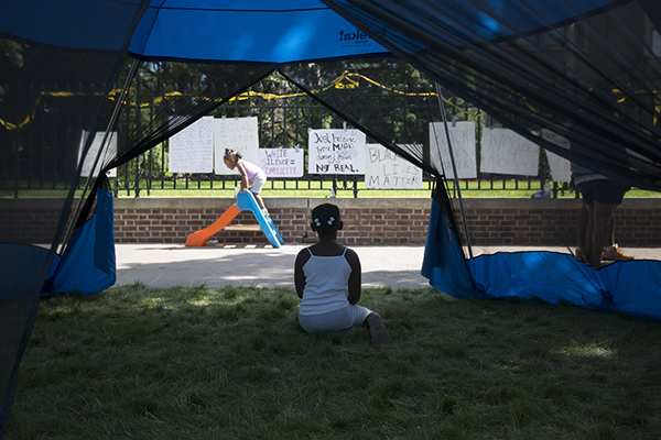 A child sits on the grass in front of a large fence covered in Black Lives Matter protest signs, another child plays on a small toy slide on the sidewalk in front of the fence.