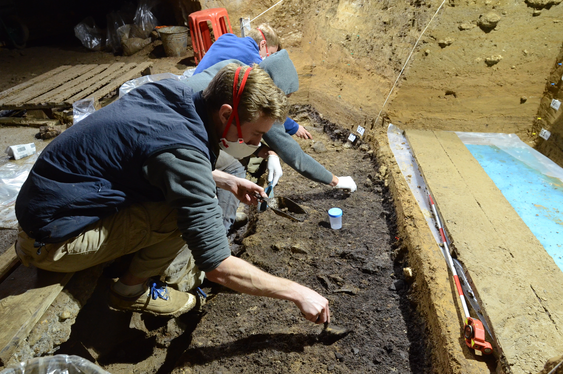 People squatting in a cave with face masks on, digging through dirt and clay. 