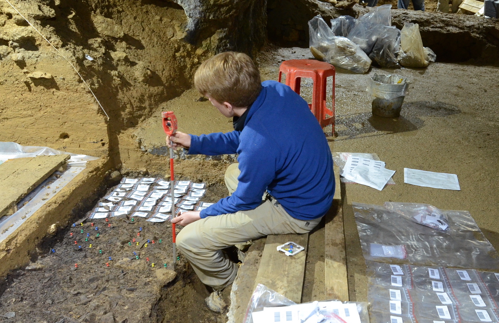 A person using a tool during an archaeological dig in a cave. In front, on dirt, sit about 30 samples placed on top of small white sheets of paper. Behind are plastic bags to hold the findings.