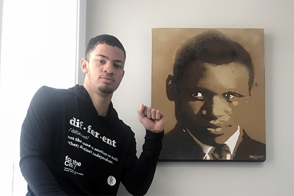 Jelani Williams holds up a fist of solidarity while seated next to a painting of singer, actor, athlete, and political activist Paul Robeson.