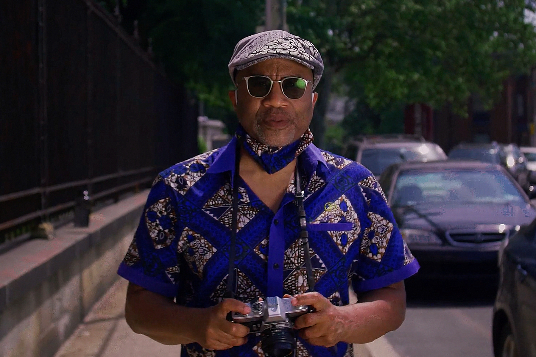 Professor Guy Ramsey standing on sidewalk with cars in street next to him, wearing a cap, sunglasses, and a mask under his chin, holding a camera.