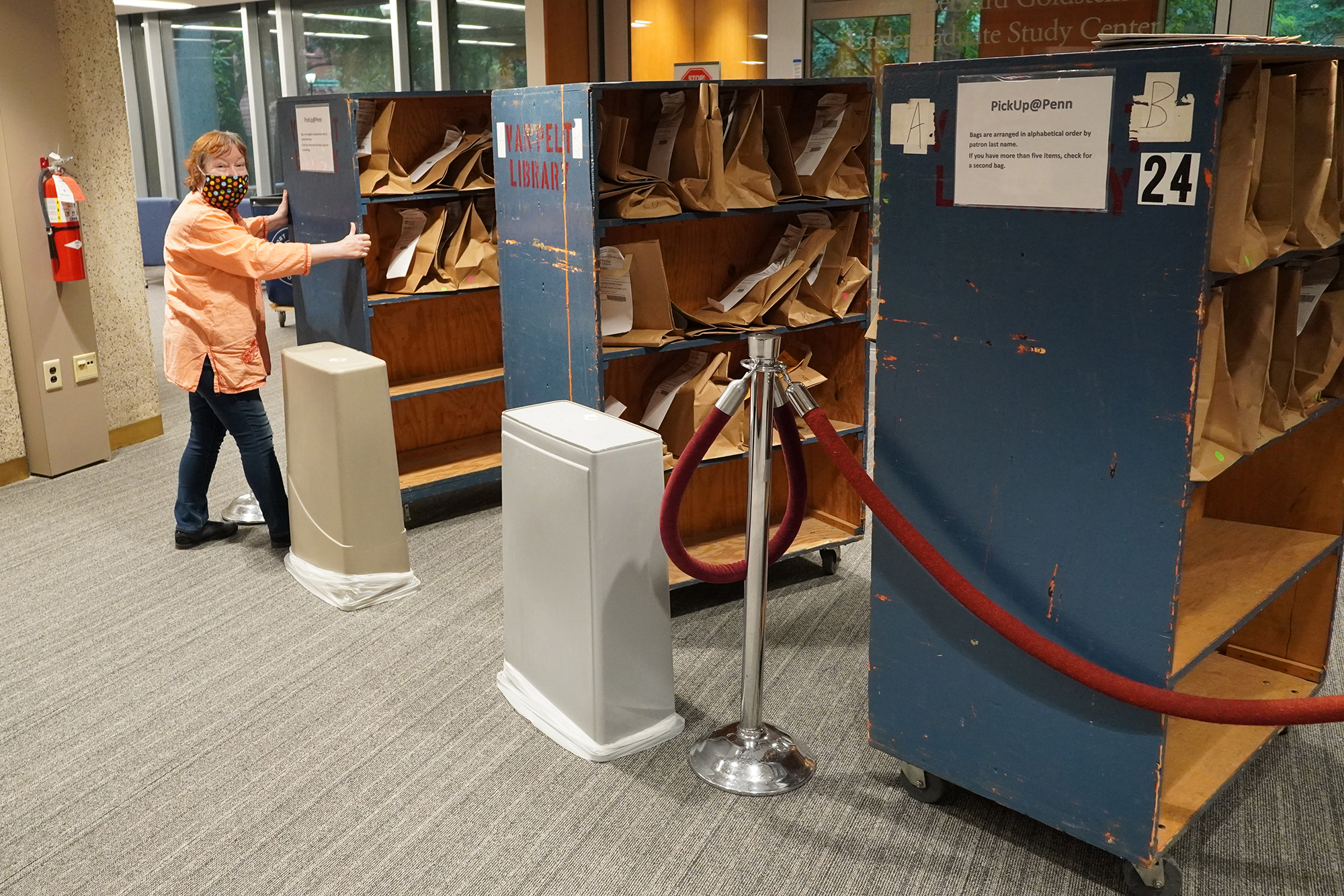 Emily Batista inside the library wearing a face mask stands by a large cart of bookshelves.