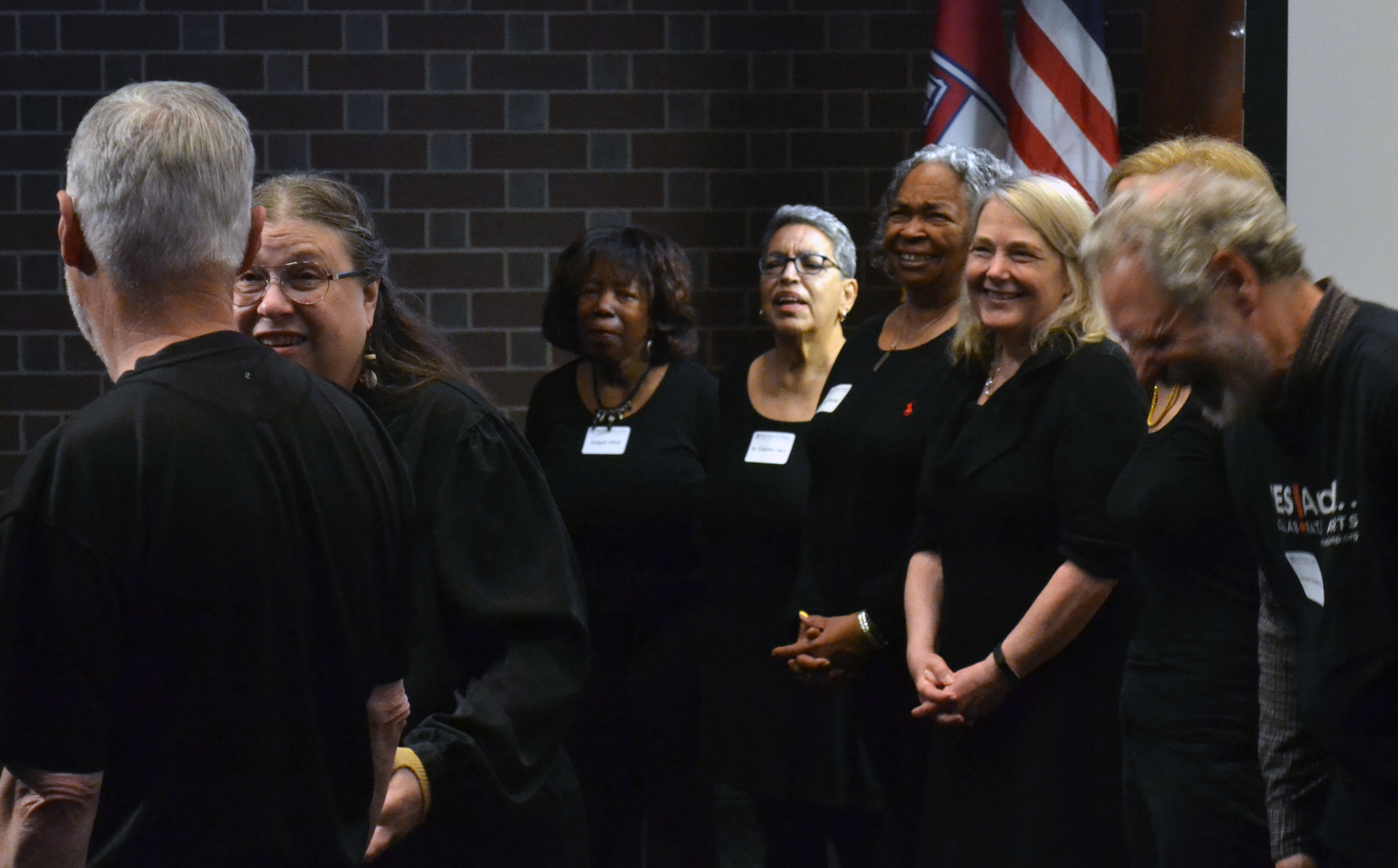 A group of people wearing all black clothing standing in a room with a brick wall and American flag in the background.