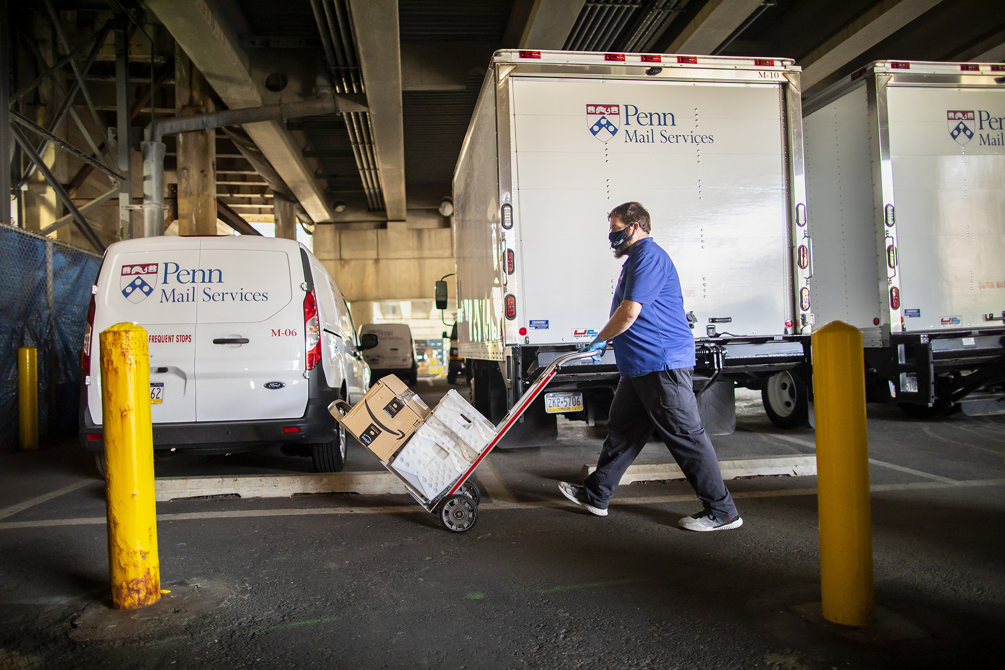 person pushes a hand truck piled with mail and packages with Penn Mail Services trucks in background. Photo by Eric Sucar.
