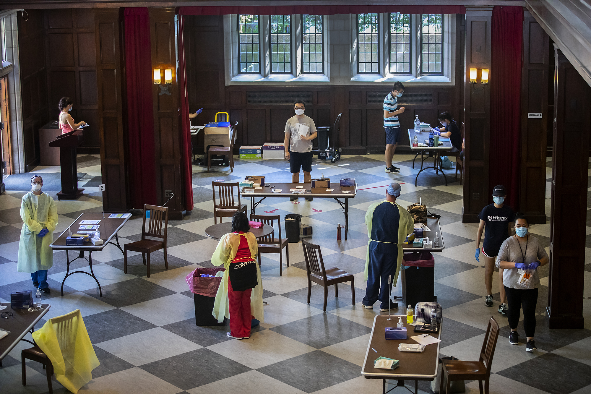 A large room in Houston Hall set up with tables and medical personnel in PPE standing by to administer tests to four masked students waiting.