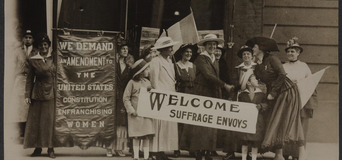 Group of women holding a banner reading Welcome Suffrage Envoys shaking hands with other women arriving at the building.