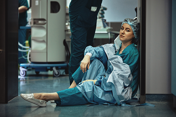 Tired nurse sitting on hospital floor in PPE with mask removed and a hospital staffer behind them.