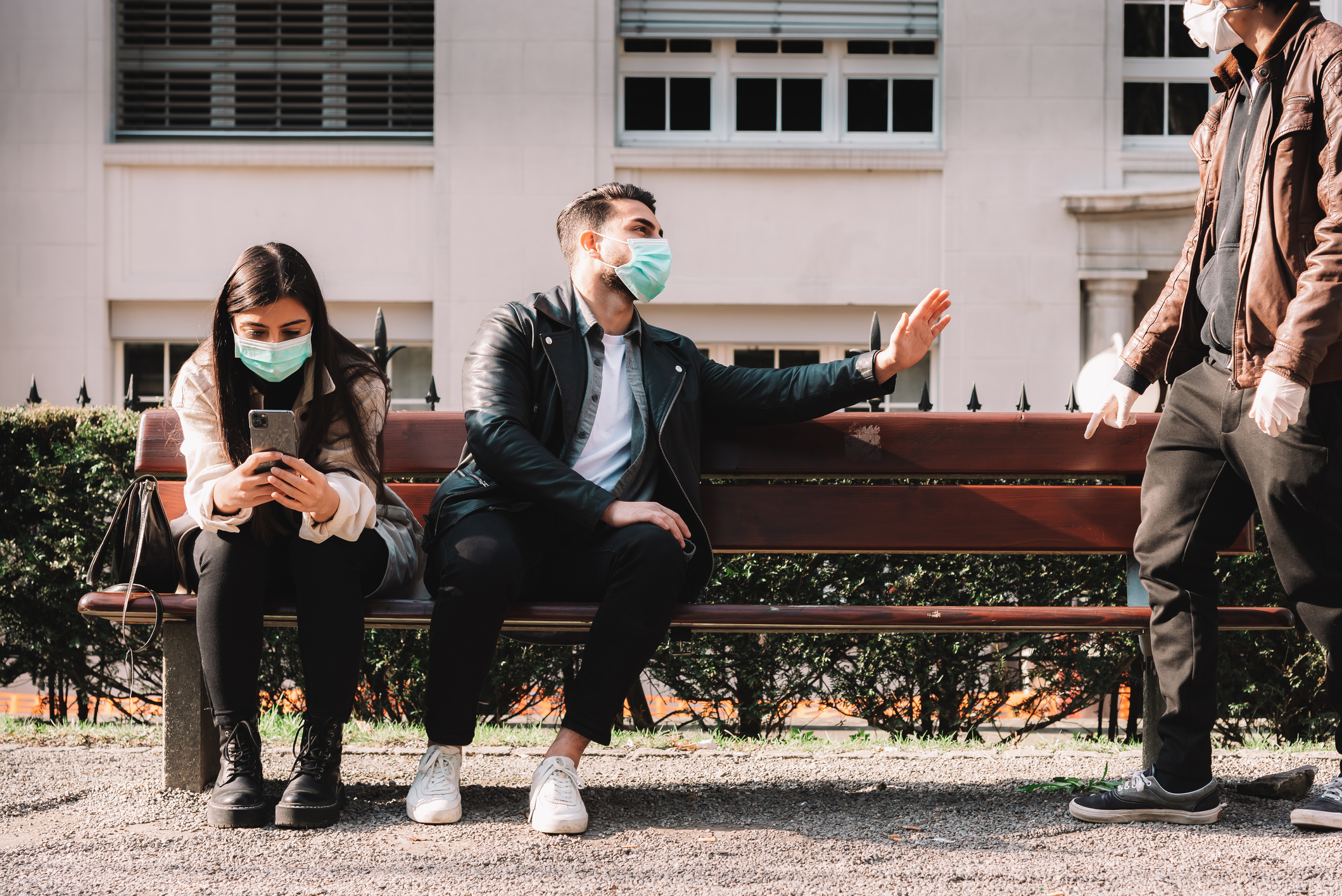 two people on a bench wearing masks, a third person motions that they want to sit in an open spot while one person currently on the bench gives a hand motion indicating no