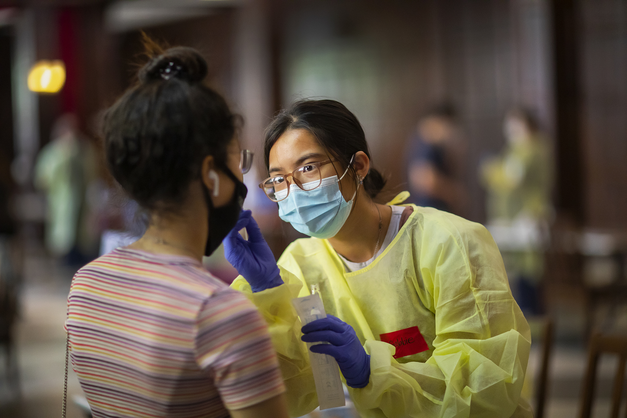 A person in a surgical gown, rubber gloves, face mask, and glasses holding hands in front of the face of a person whose back is to the camera. That person is also wearing a mask and has earbuds in.