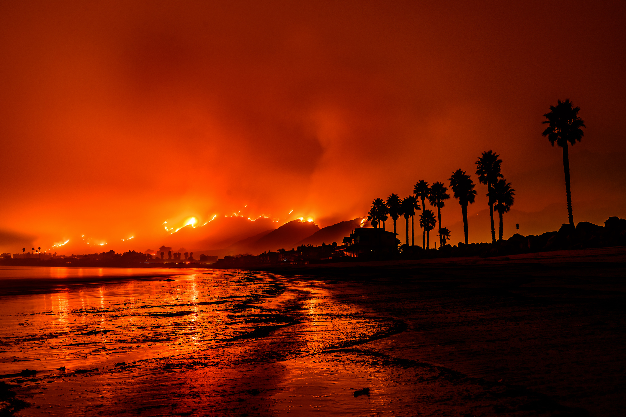 Water washes up on the shore at the beach while a fire rages in the background.