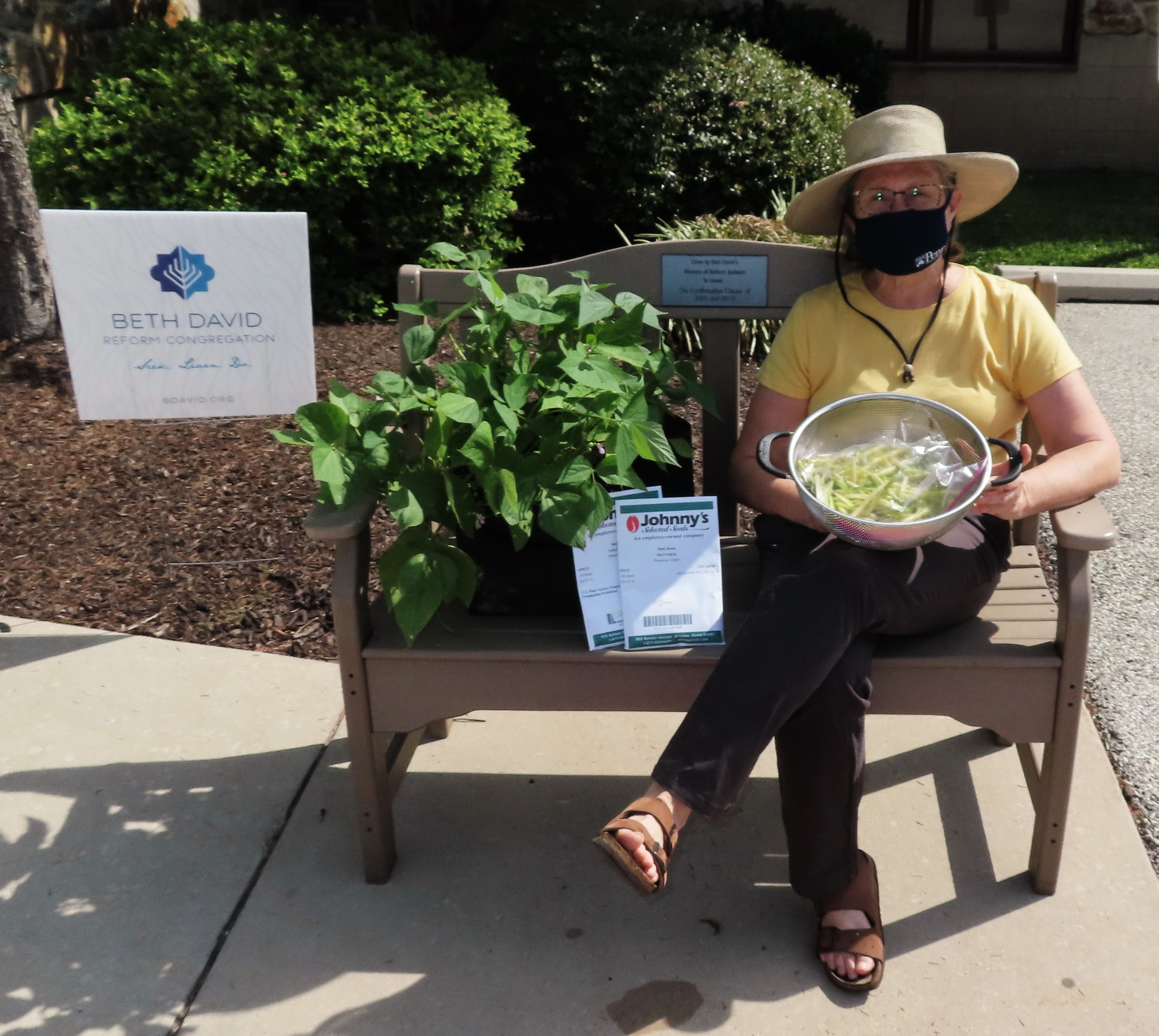 Person with a mask sitting on a bench holding a bowl of green beans next to a sign that says Beth David 