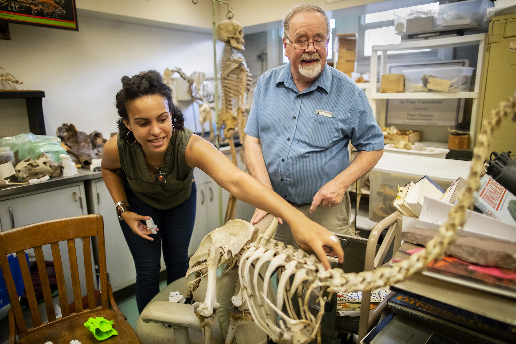 Aja Carter and Peter Dodson standing in a room full of dinosaur skeletons and books, Carter is touching the vertebrae of a skeleton.
