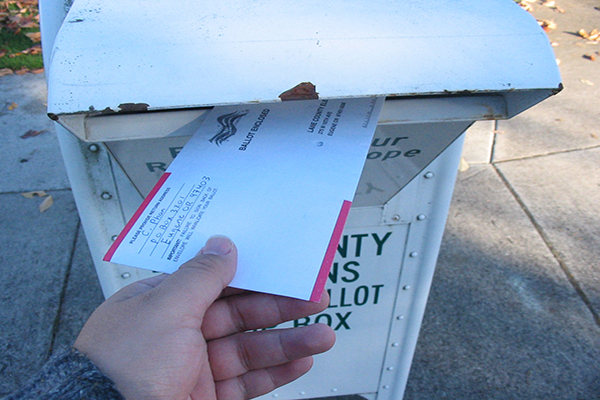 A hand can be seen putting an envelope with the words "ballot enclosed" into a ballot box on a sidewalk.