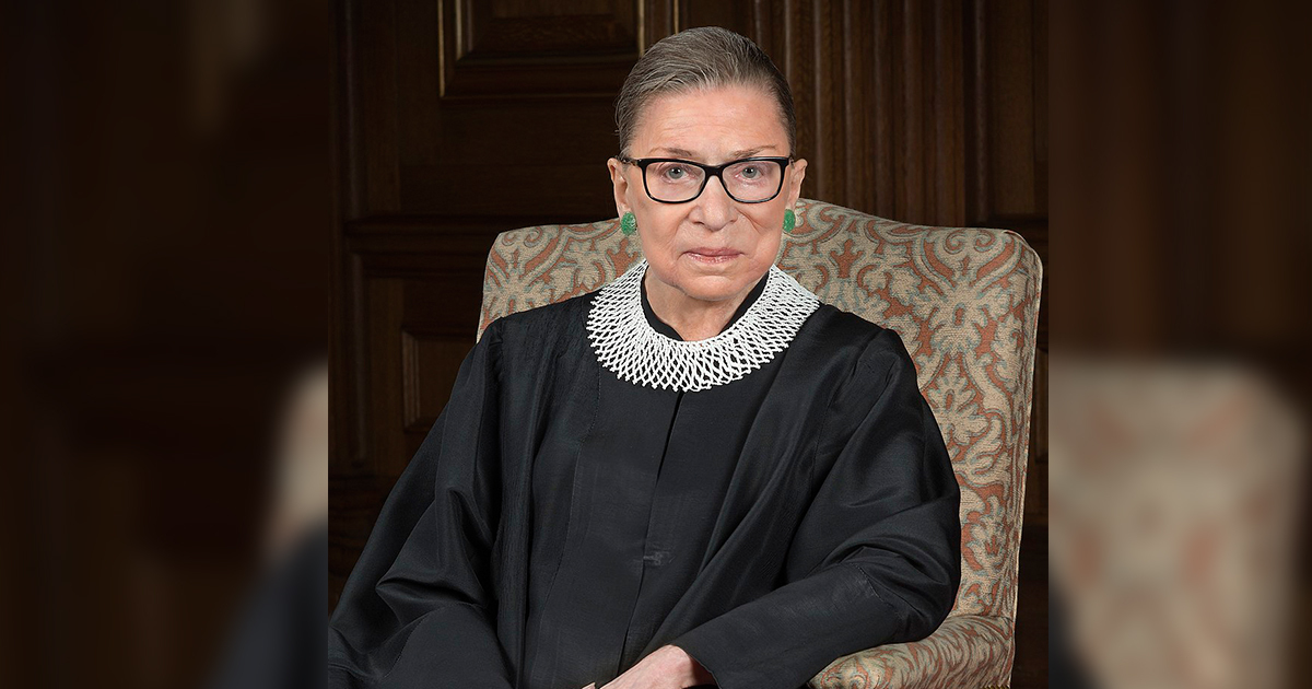 Person with hair pulled back, green earrings and black glasses looks into the camera, wearing Supreme Court robes and white lace collar.
