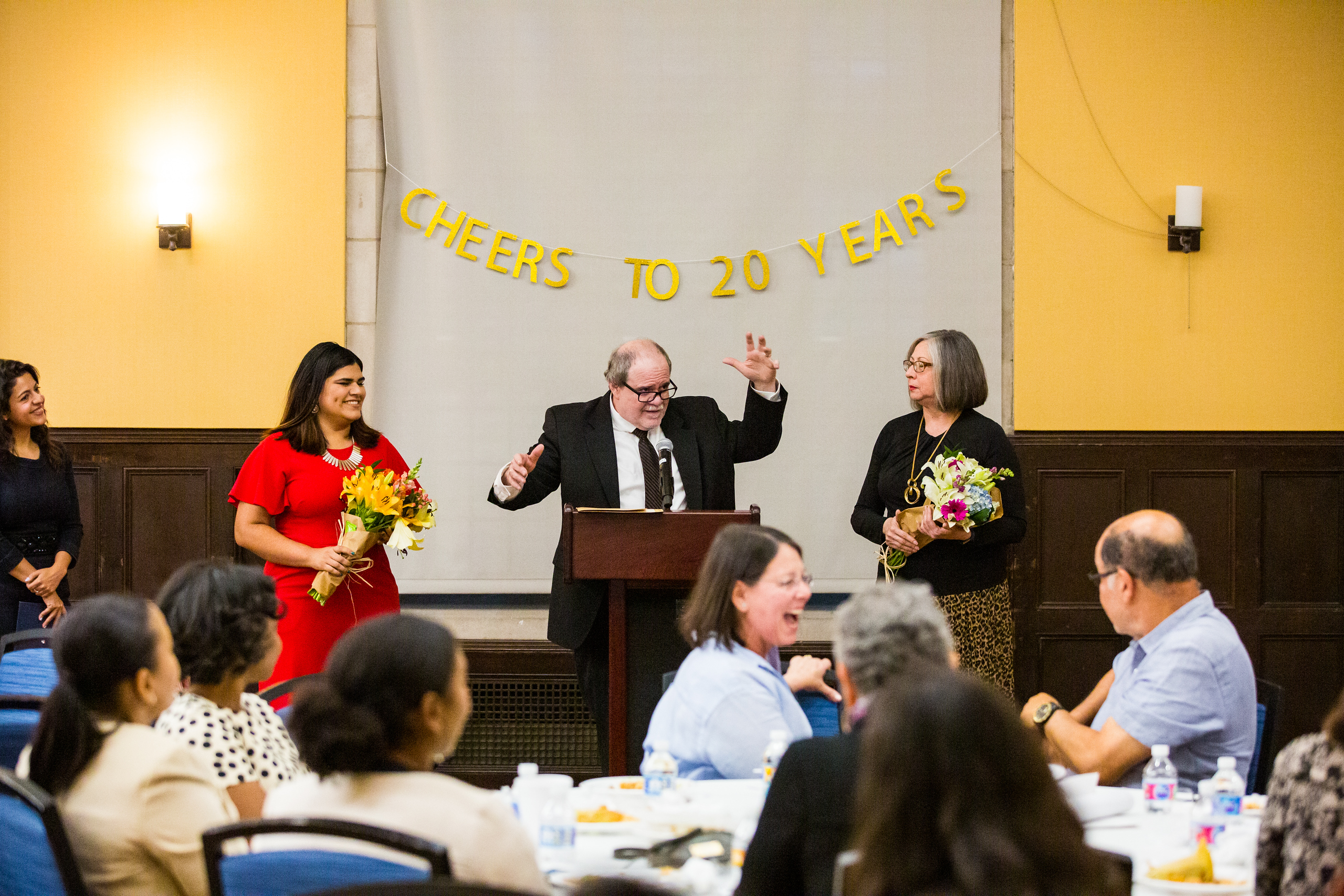 A banner reading "cheers to 20 years" hangs behind a man (center) and three women at a podium