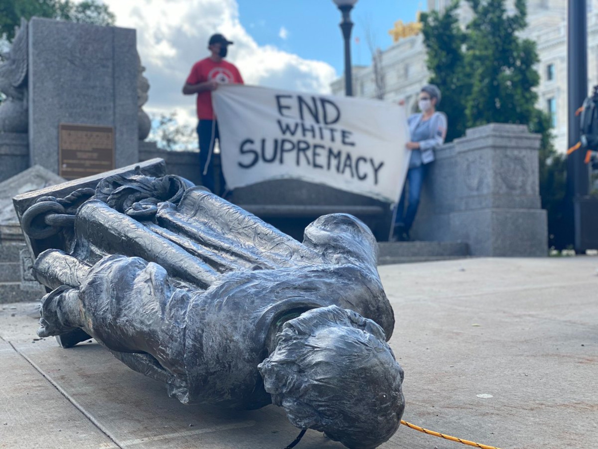 A toppled statue of Columbus near the Minnesota State Capitol. Two people hold an End White Supremacy sign in the background.