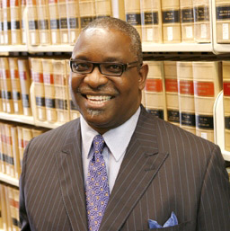 Man in glasses and suit and tie smiles as he stands in front of rows of law books on library shelves.