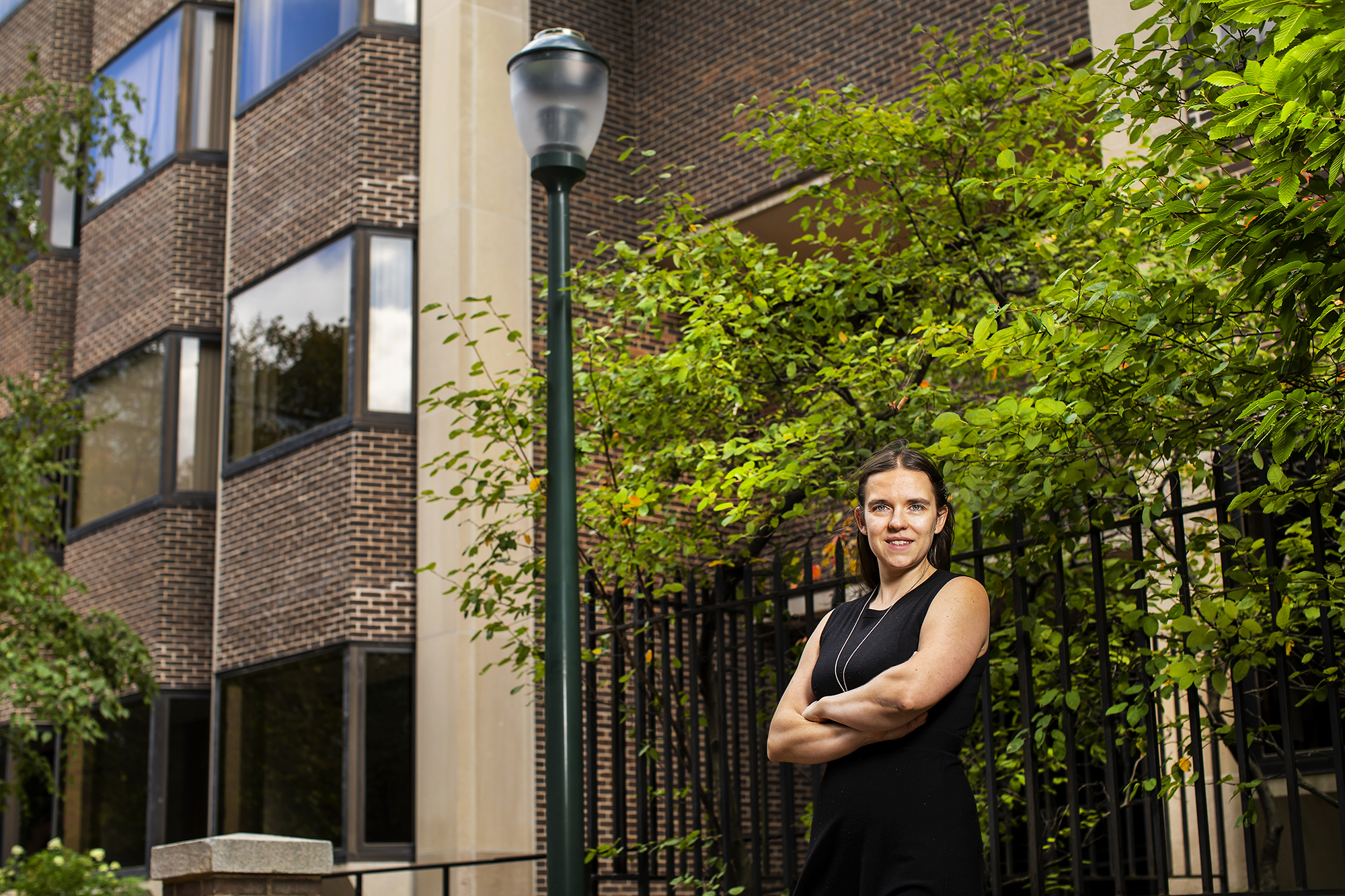 Person standing outside of a brick building, in front of a lamppost and shrubbery, arms crossed.